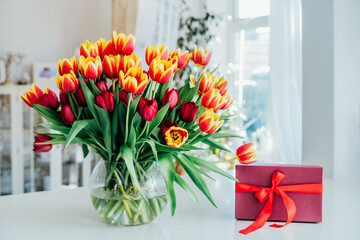 Huge spring bouquet of tulips in vase and gift box on the table with light classic interior design background. Gift for holiday, birthday, 8 March, Mother's Day, Valentine's day, Women's Day.