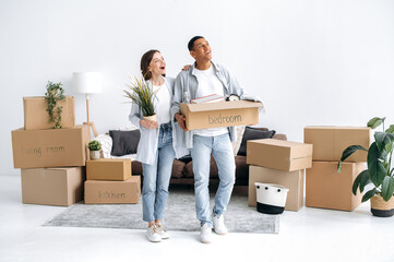 Moving to a new home. Excited couple in love, husband and wife of different nationalities, moved to their new home, guy holds a box with things, girl a flowerpot, enthusiastically inspecting new house