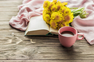 Morning coffee. A bouquet of yellow dandelions on an open book and a cup of coffee on a wooden table. Cozy morning. Copy space.
