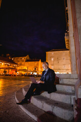 Geschäftsmann sitzt nachts auf Stein-Treppe und arbeitet am Laptop, Platz mit alten Häusern, Hochformat