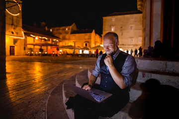 eleganter Mann sitzt nachts auf Stein-Treppe und arbeitet nachdenklich am Laptop, Platz mit alten Häusern, Hochformat