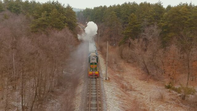 Along beautiful forest, country road. Flying above retro tourist train crossing picturesque countryside. Vintage vehicle passing through snowy countryside. Aerial view heritage steam train in woodland