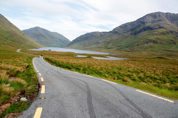 Small narrow road into mountains by a lake. Car tire print of asphalt surface. Boy racers in rural area. Beautiful landscape of Connemara. Ireland.
