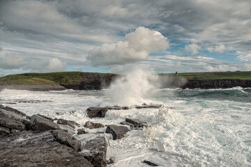 Powerful ocean wave crushes on rock, cliff with green fields and blue cloudy sky in the background. Beautiful nature scenery. Doolin, county Clare, Ireland. Irish landscape.