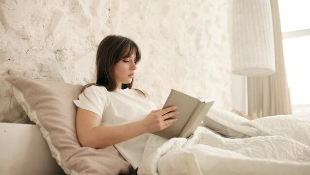 Portrait of a calm attractive young woman reading a book in the bed in the morning. Calm morning concept. High quality 4k footage