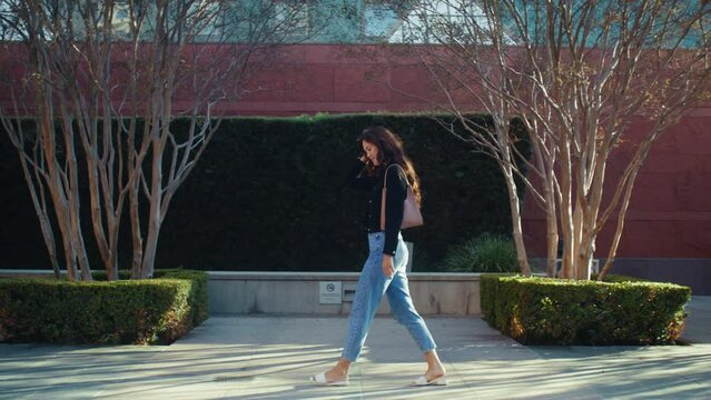 Relaxed brunette walking street near green plants. Asian girl going on pavement.