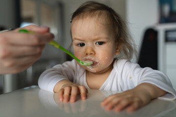One baby close up on small caucasian girl and hand of her unknown mother feeding her at home in day small child with food around mouth making mess dirty while eating real people growing up copy space
