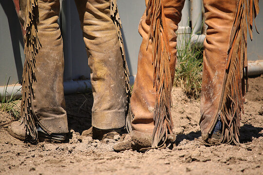 Western lifestyle rearview image of the lower legs of two cowboys wearing leather chaps and cowboys boots covered in mud.