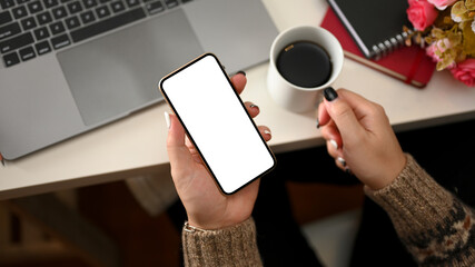 Female in cozy and warm sweater holding a smartphone over her working desk.