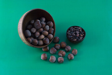 Hazelnuts are scattered from a large wooden bowl. Pine nuts in a small cup. Green background