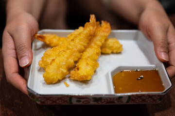 Close up hands holding fried shrimps tempura with chili sauce on white dish and wood table background.
