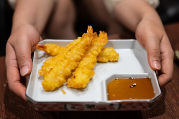 Close up hands holding fried shrimps tempura with chili sauce on white dish and wood table background.