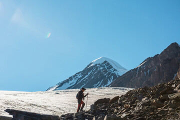 Scenic alpine landscape with silhouette of hiker with trekking poles against large glacier tongue and snow mountain peak in sunlight. Man with backpack in high mountains under blue sky in sunny day.
