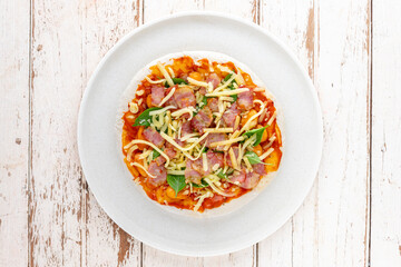 raw pizza with tomato sauce, sausage, bacon, sweet basil, oregano, mozzarella, cheddar, gouda, romano cheese in white ceramic plate on white wood texture background, top table view, flat lay
