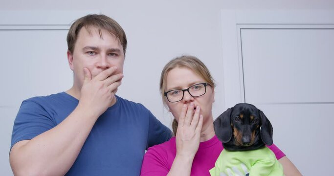 Young couple smiles happily, but suddenly sees or remembers something bad, so their facial expressions change and they cover mouths with hands in shock. Woman holds cute dachshund puppy in her arms