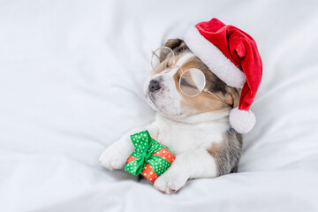 Tiny Beagle puppy wearing red santa hat sleeps and hugs gift box under white blanket at home. Top down view. Empty space for text