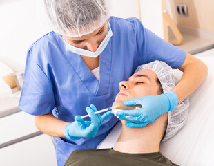 Professional female beautician performing face contouring injections to male patient at cosmetology...
