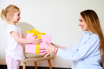 Happy women's day. Little girl is giving her young mom present, woman is smiling while sitting on floor at home. Happy family with Christmas or birthday gift