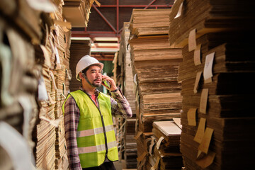 Asian male worker in safety uniform and hard hat, supervisor inspector talks with mobile phone about packaging stock order at crate factory with piles of stacking paper, recycling production industry.