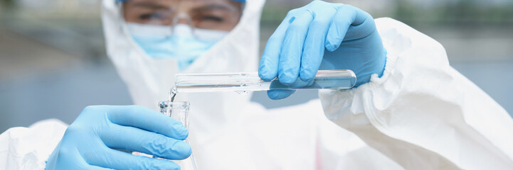 Researcher in protective suit and glasses with gloves pours water from test tube glass beaker