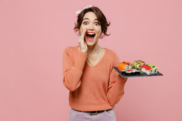 Young woman in sweater hold makizushi sushi roll served on black plate traditional japanese food scream hot news about sales discount with hands near mouth isolated on plain pastel pink background