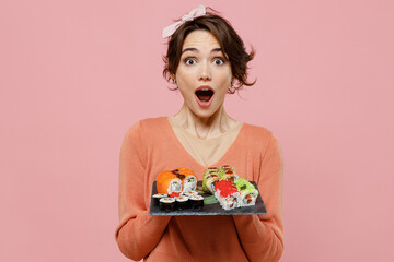 Young amazed woman 20s wear casual clothes hold in hand makizushi sushi roll served on black plate traditional japanese food look camera with opened mouth isolated on plain pastel pink background