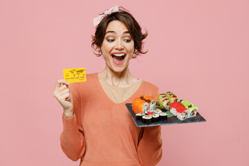 Young amazed happy woman 20s in casual clothes hold in hand makizushi sushi roll served on black plate traditional japanese food hold in hand credit bank card isolated on plain pastel pink background.