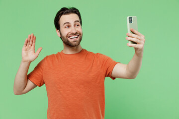 Young fun man 20s wear orange t-shirt doing selfie shot on mobile cell phone post photo on social network waving hand say hello isolated on plain pastel light green color background studio portrait
