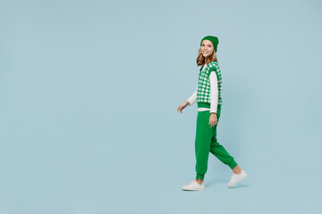Full size body length side view profile fun excited stunning young brunette girl teen student wear checkered green vest hat go move walk isolated on plain pastel light blue background studio portrait