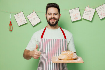 Young smiling fun male chef confectioner baker man 20s in striped apron hold warm homemade bread look camera show thumb up isolated on plain pastel light green background studio Cooking food concept