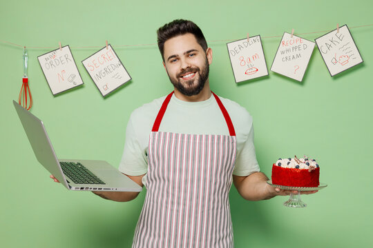 Young satisfied fun male chef confectioner baker man in striped apron hold cake use work on laptop pc computer look aside isolated on plain pastel light green background studio. Cooking food concept.