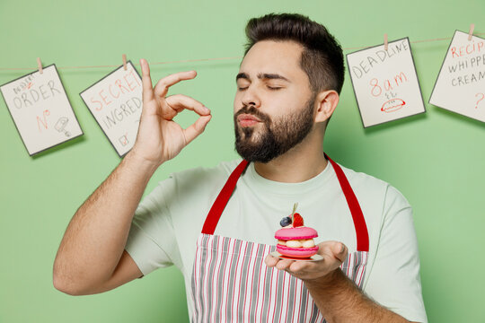 Young male chef confectioner baker man in striped apron holding pink little cake muffin macaroon show belissimo perfect gesture isolated on plain pastel light green background Cooking food concept.
