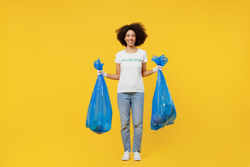 Full body young woman of African American ethnicity in white volunteer t-shirt hold trash bags after rubbish removing isolated on plain yellow background. Voluntary free work assistance help concept