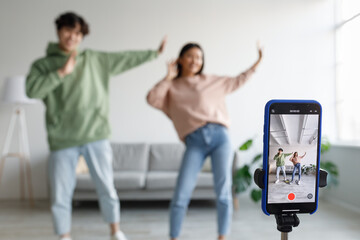 Millennial Asian couple recording video, having fun, dancing on mobile phone camera at home, selective focus