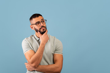 Pensive middle eastern guy thinking and touching chin, looking aside at free space, posing over...