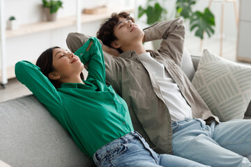 Peaceful young Asian couple relaxing on sofa with their hands behind heads in living room