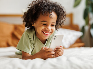 Cute little african american boy with smartphone watching cartoons or playing mobile games at home