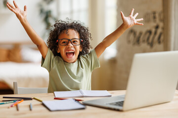Little african american school boy raising hands up with excitement during home distance education - 486209250