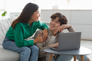 Happy millennial Asian couple using laptop, surfing web, watching movie, relaxing at home on weekend morning