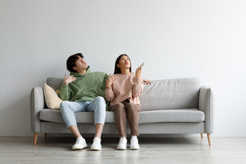 Young Asian couple suffering from summer heat, holding remote control, switching on air conditioner at home, mockup