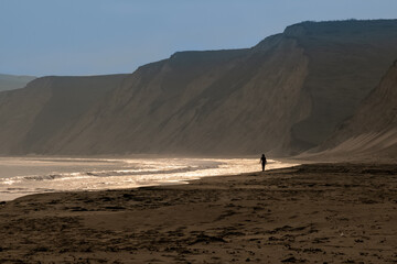  A solitary figure in silhouette walks beneath the cliffs on the shoreline of a beach in the glow...