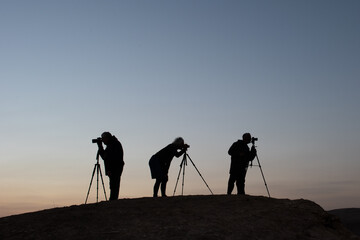 Silhouette of 3 photographers standing next to tripod and camera at sunrise on a desert hilltop. 