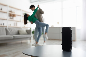 Millennial Asian couple dancing to popular music at home, selective focus on portable wireless speaker on table