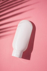 Plastic white tube for cream or lotion. Skin care or sunscreen cosmetic in top view on pink background with palm leaves shadow. Beauty concept for face care