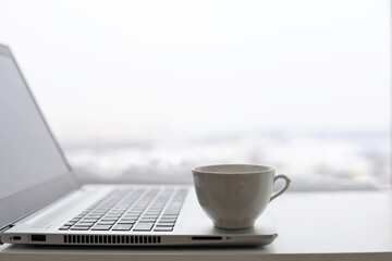 Coffee cup and laptop on table against the window, view to winter city. Cozy workplace in home office