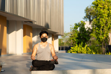 asian man short hair wear black face mask, plant sneaker and grey vest do meditation in park near modern building and green graden evening morning light