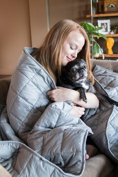 Young Redhead Woman And Her Dog Using Soft Weighted Blanket In Her Modern Apartment With Her Pet Dog