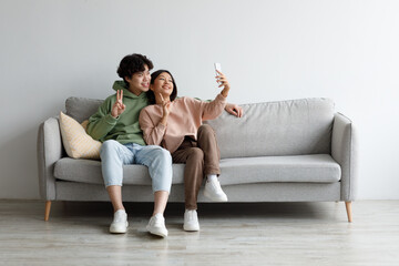 Obraz premium Portrait of happy millennial Asian couple taking selfie together, sitting on sofa at home, copy space