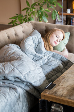 Young Redhead Woman And Her Dog Using Soft Weighted Blanket In Her Modern Apartment While Sleeping On Couch