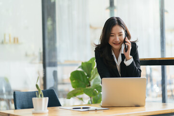 Happy smiling Asian business woman working on laptop at office, using smart phone. Businesswoman...